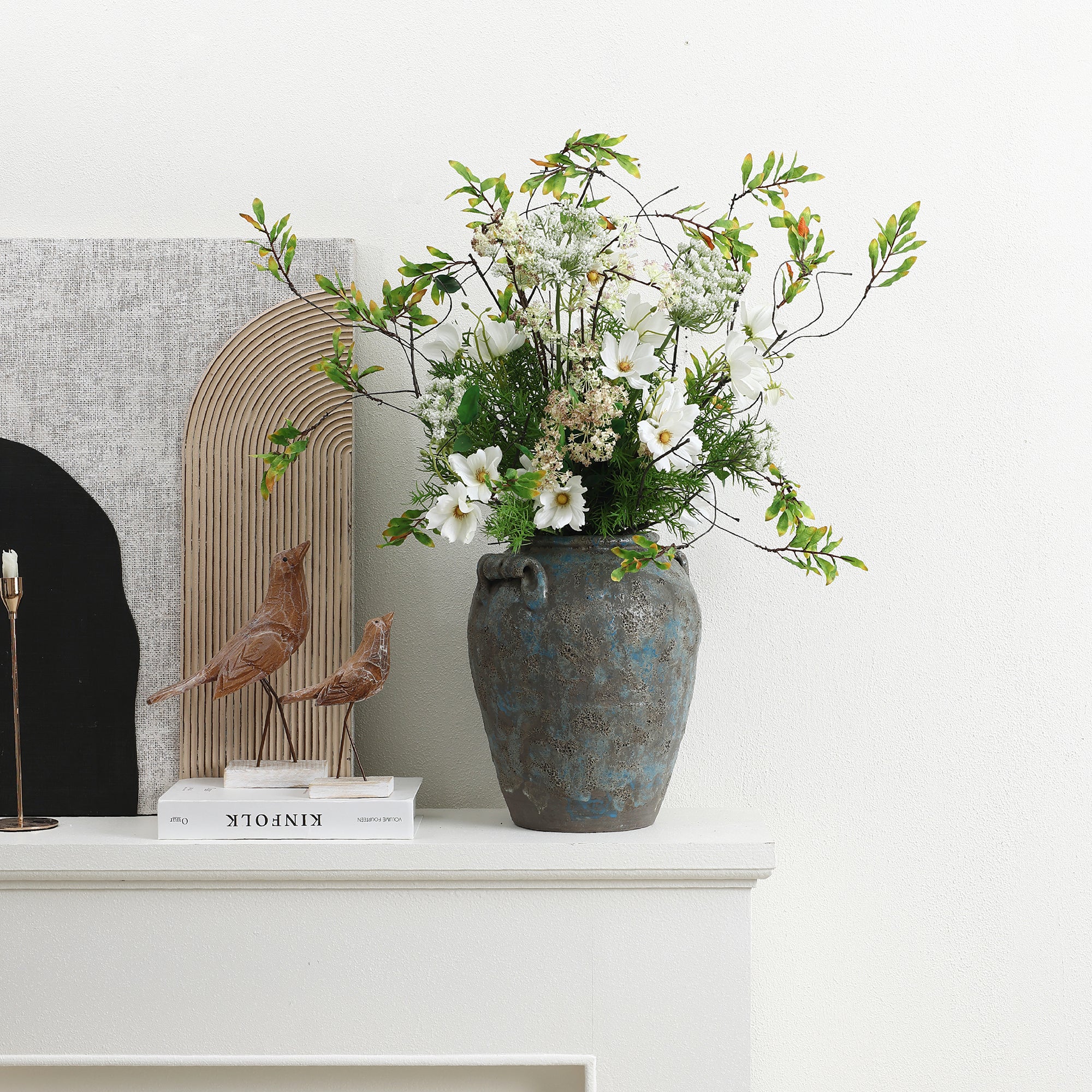Terracotta vase with white flowers and greenery sits on a white mantel beside books, wooden bird figurines.