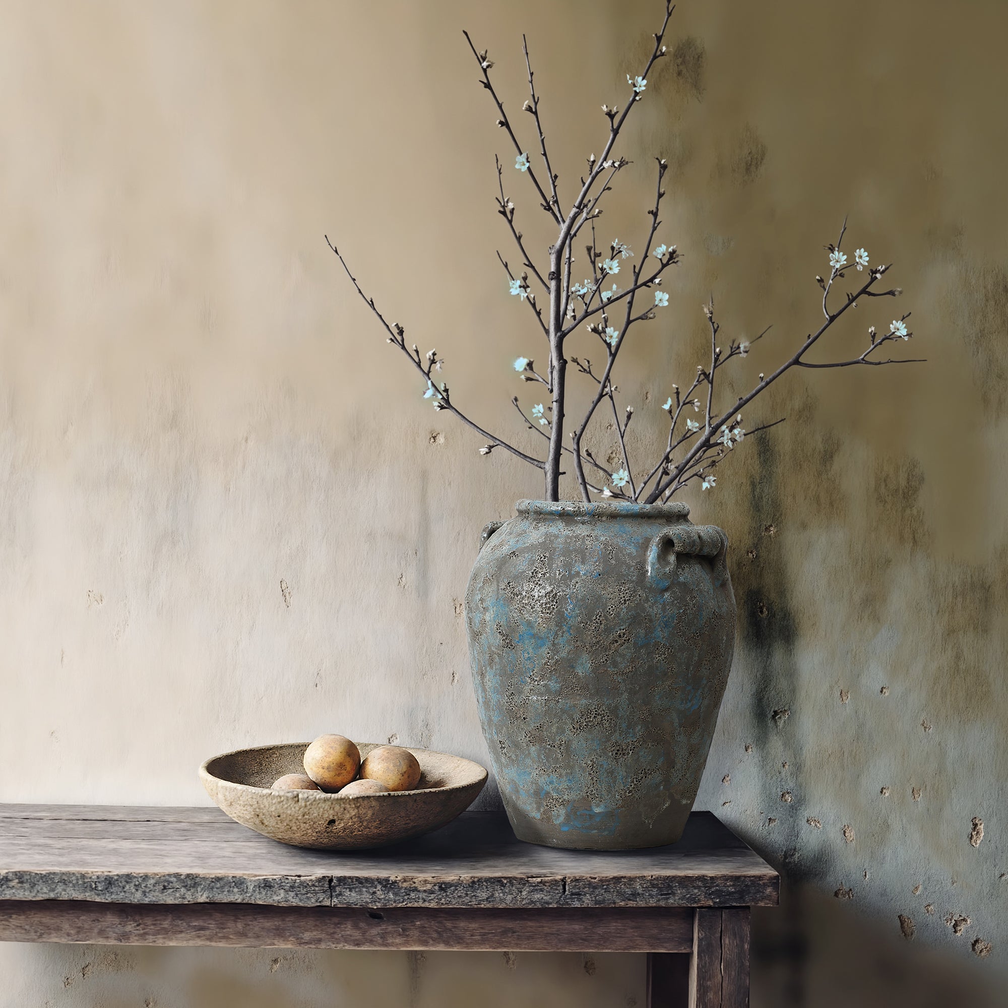 Terracotta vase with blooming branches and a shallow bowl of three round fruits sit on a weathered wooden table.