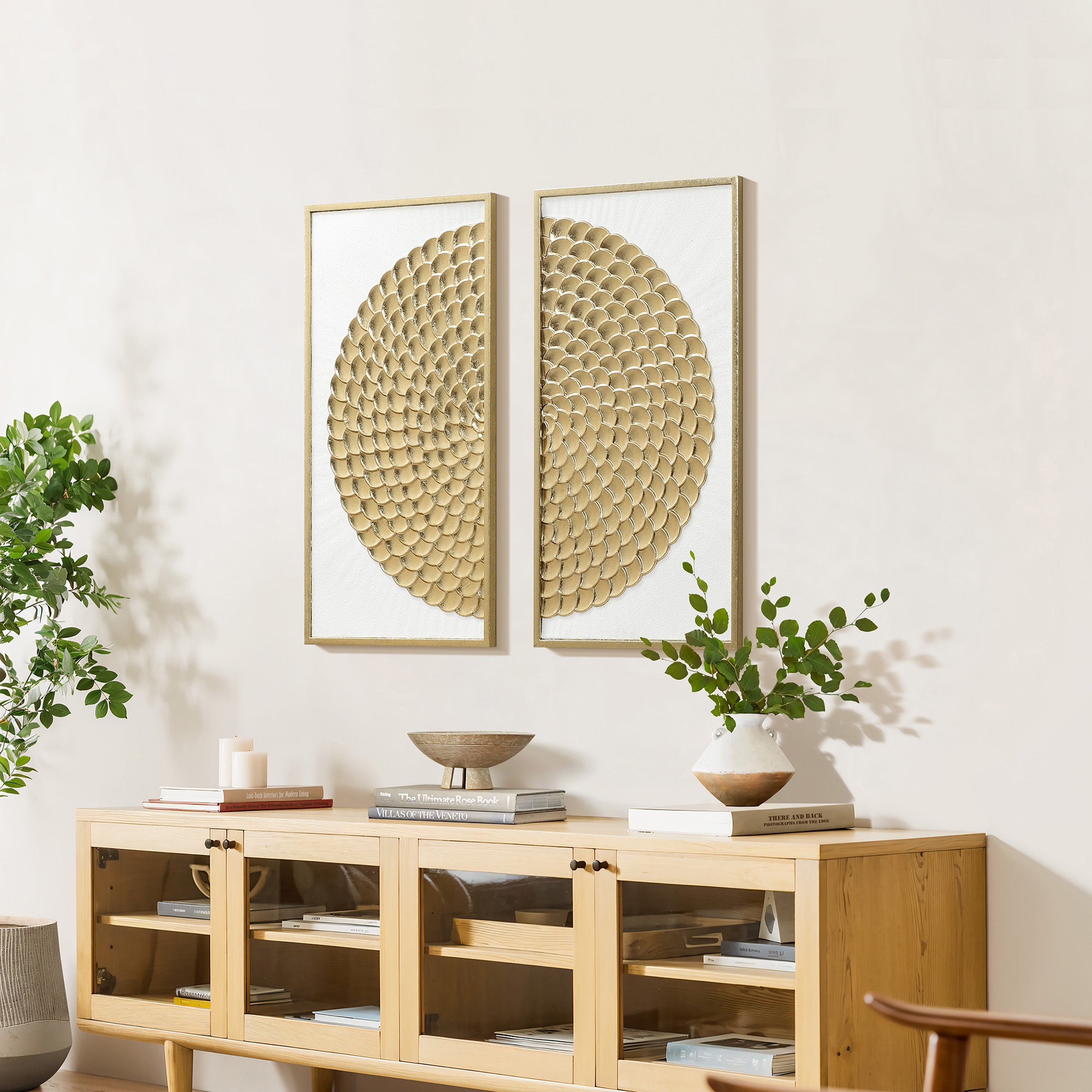 Two white and gold wall art are displayed above a  cabinet with decorative vases, a bowl, and a woven chair in the foreground.