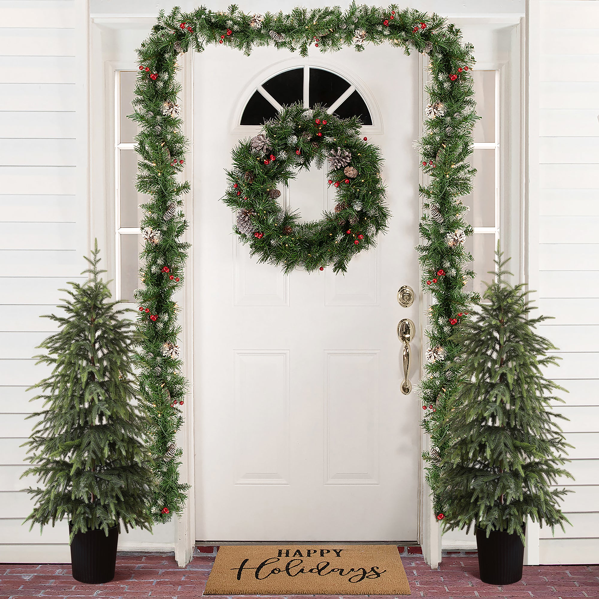 White front door is decor with wreath,garland red ornaments, flanked by 2 potted artificial trees ,doormat reading Happy Holidays.