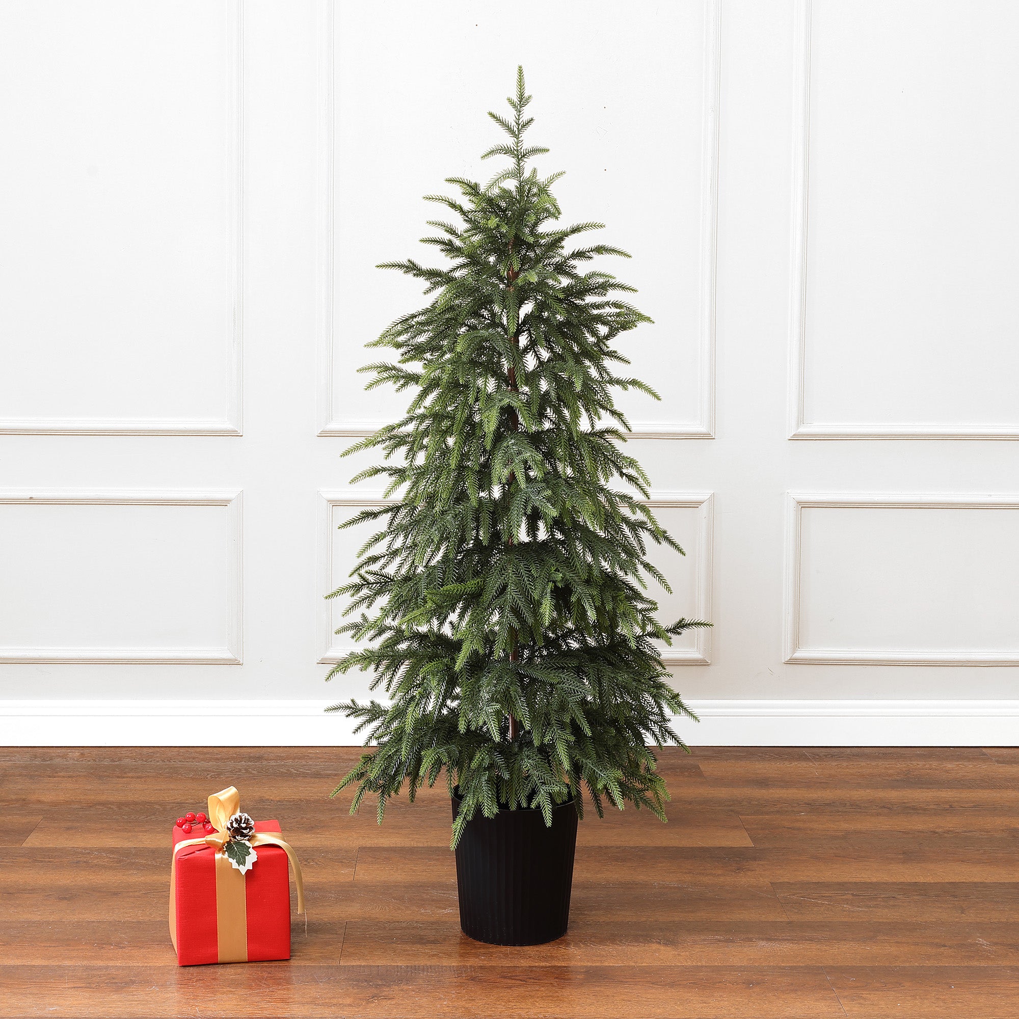 Slim artificial tree stands on wooden floor beside single red gift box with gold ribbon, in front of white paneled wall.