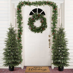 White front door decor with wreath, garland is flanked by 2 4ft artificial christmas fir trees, a doormat reading “Happy Holidays.”.