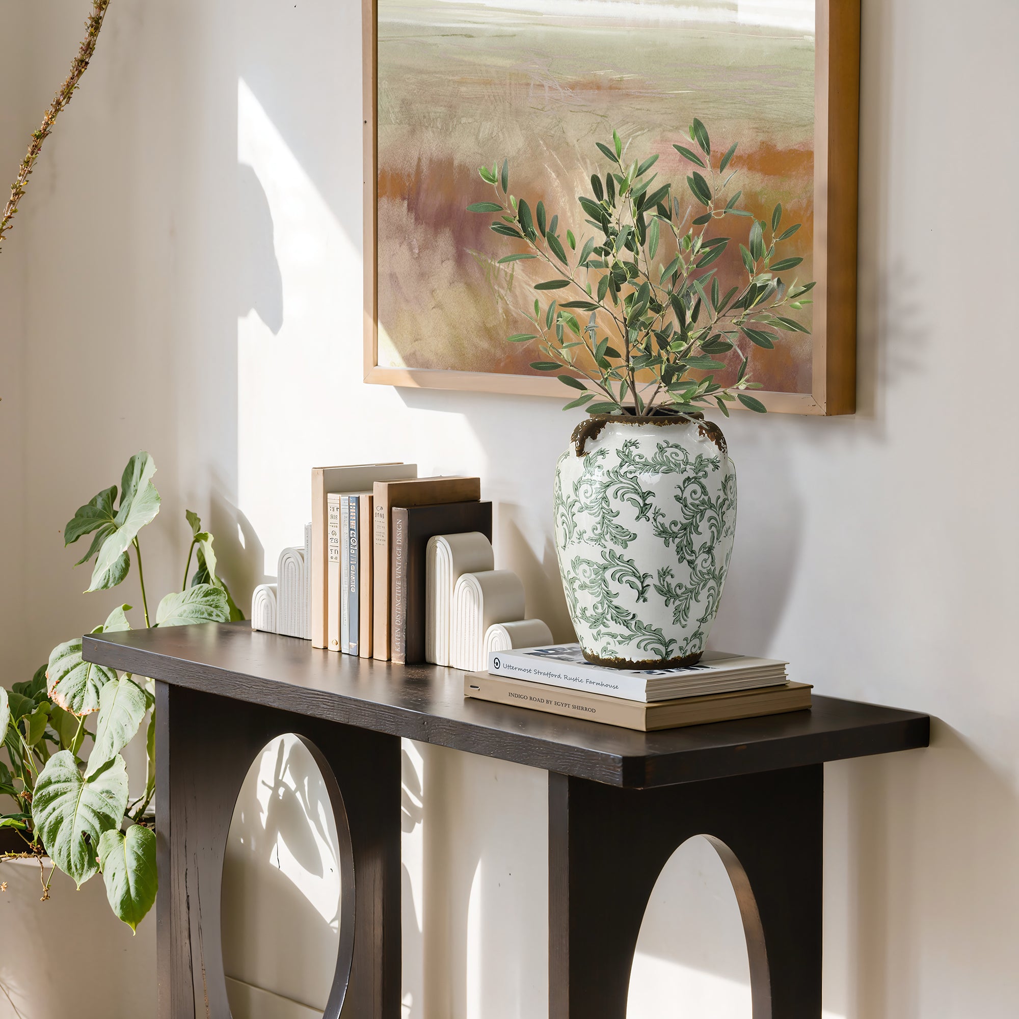 A decorative console table with books, white bookends, and a round floral ceramic vase with foliage.