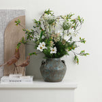Rustic ceramic vase with white and green flowers sits on a white shelf next to two decorative bird sculptures.