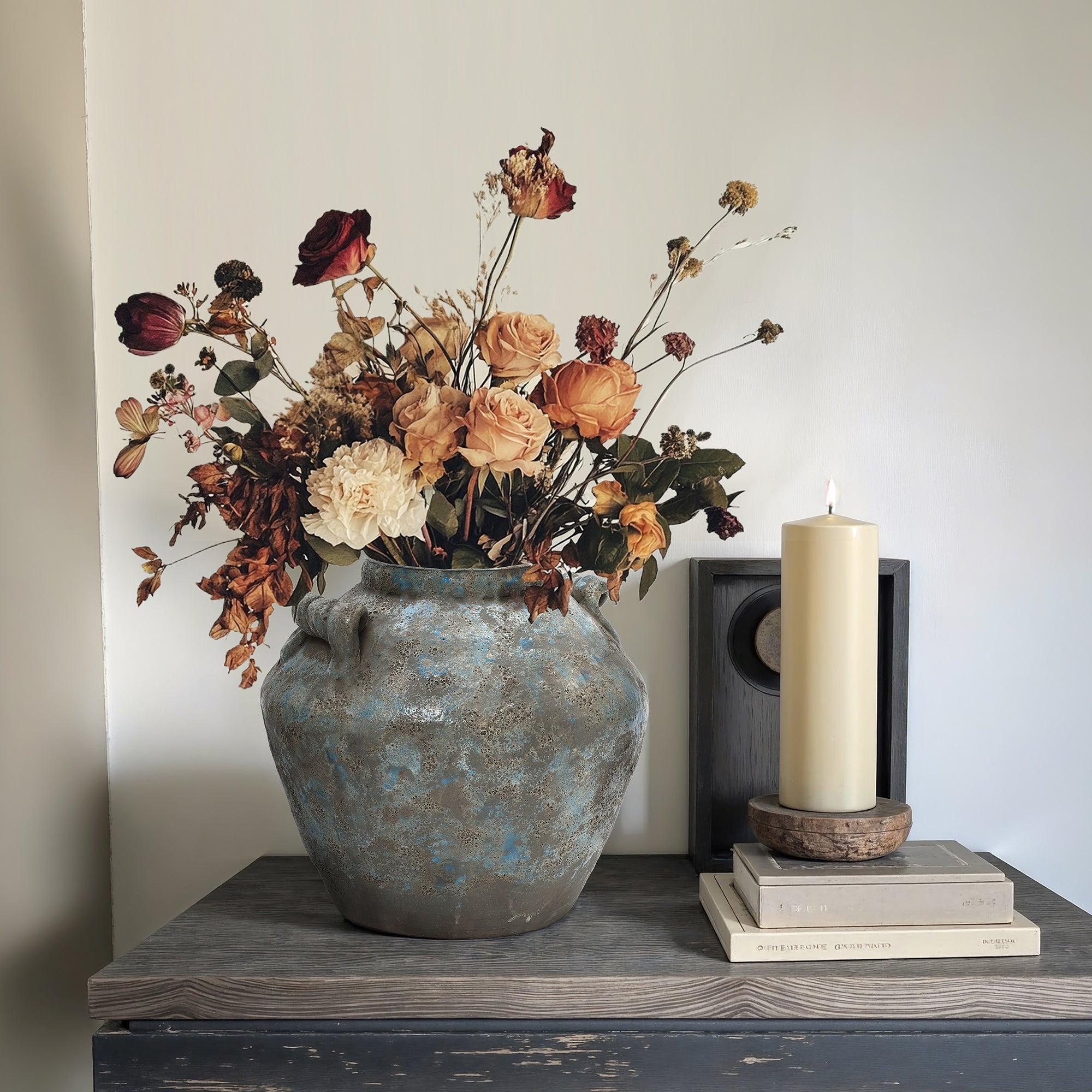 Handcrafted pottery vase with dried flowers sits on a wooden table next to a tall, lit candle resting on a stack of books.