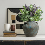 Large terracotta vase with purple and green flowers sits on a table next to two cups, abstract framed artwork.