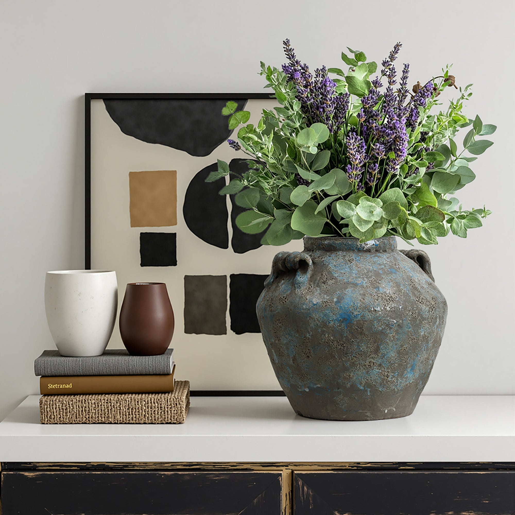 Large terracotta vase with purple and green flowers sits on a table next to two cups, abstract framed artwork.