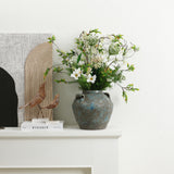 Handcrafted pottery vase with white and green flowers sits on a white shelf next to decorative objects.