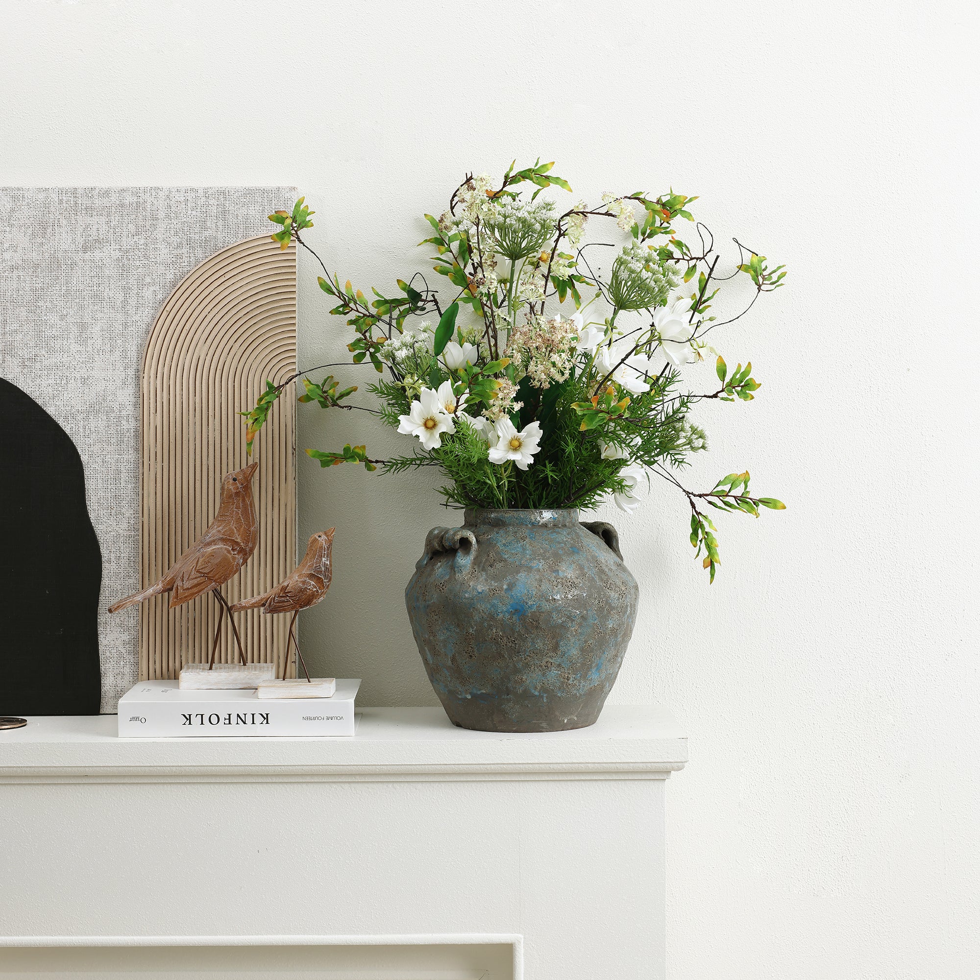 Handcrafted pottery vase with white and green flowers sits on a white shelf next to decorative objects.