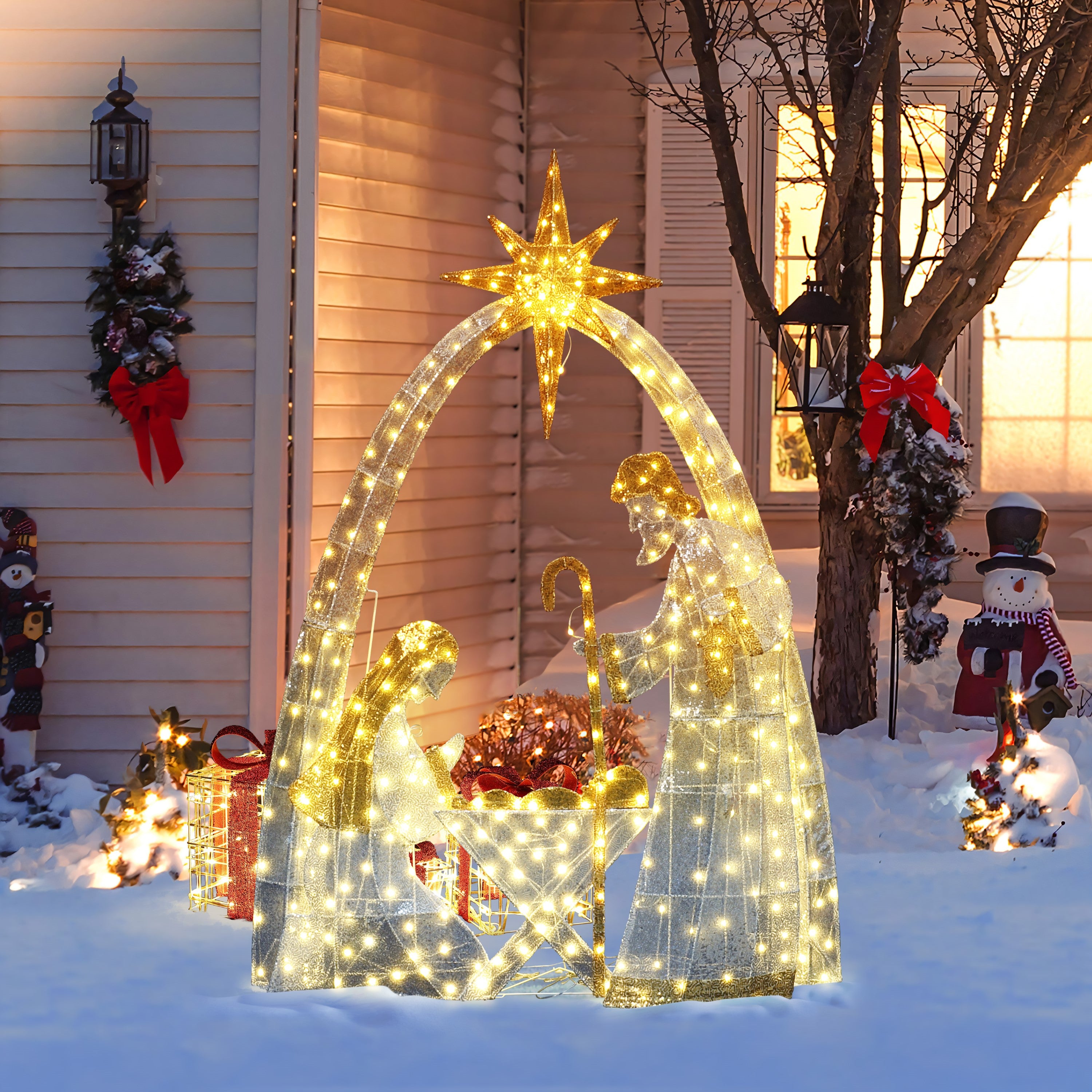 Pre-lit nativity scene with Mary, Joseph, baby Jesus is displayed on snow in front yard , surrounded by christmas decor, cheerful snowman.