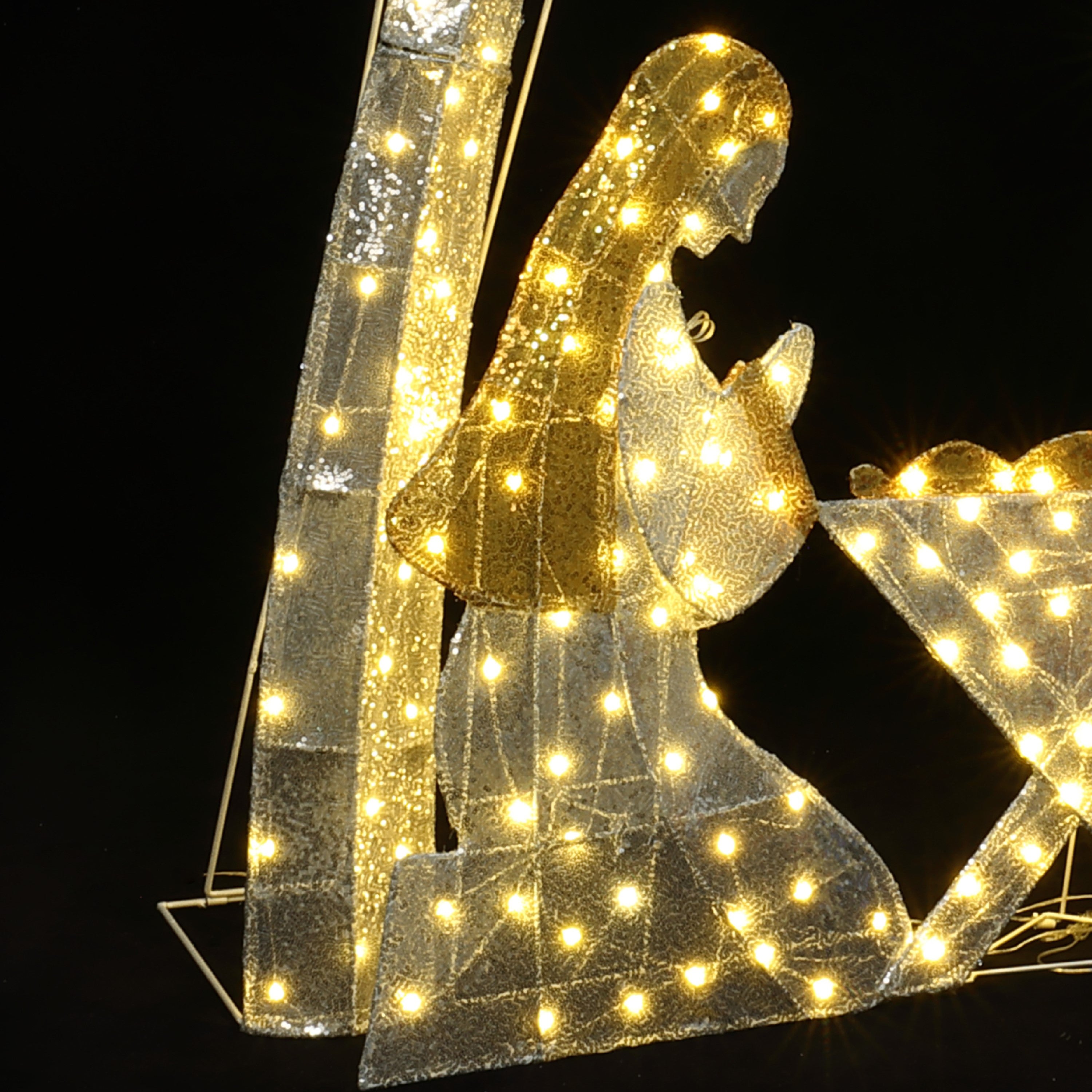 Silhouette nativity scene features Mary baby Jesus, outlined with white lights against dark background.