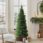 Green artificial christmas tree in wicker basket stands a chair, with wrapped gifts underneath, decor in living room with large windows ,wintry scenery outside.