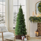 Green artificial christmas tree in wicker basket stands a chair, with wrapped gifts underneath, decor in living room with large windows ,wintry scenery outside.