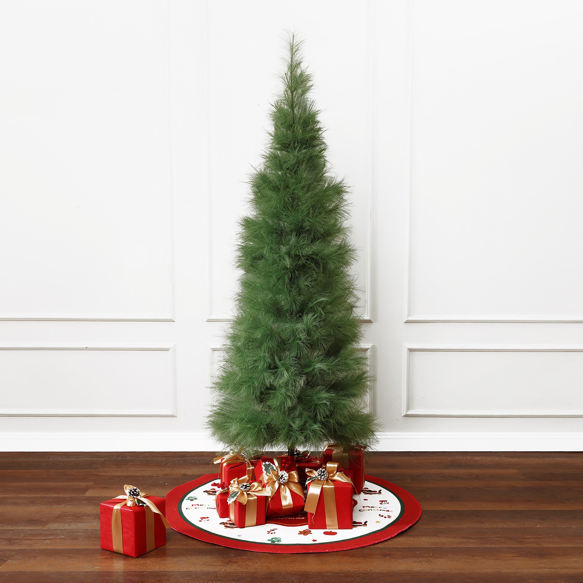 Slim 6ft pampas grass artificial christmas tree stands on white, red tree skirt with wrapped gifts beneath, against white paneled wall.