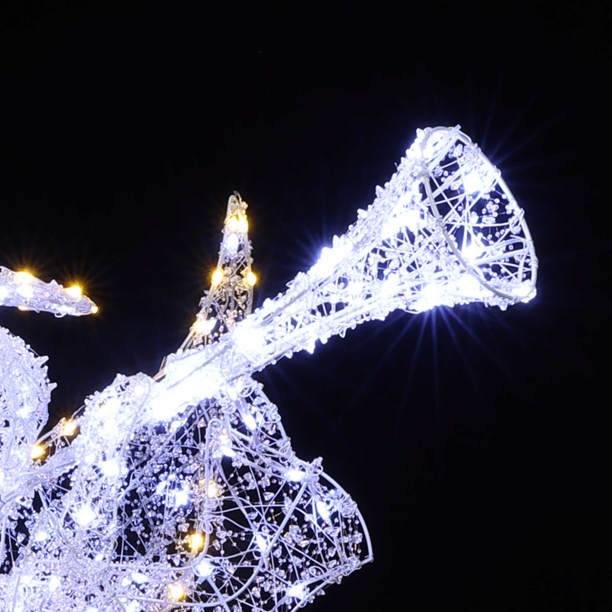 Wireframe christmas angel decor plays a trumpet, illuminated by white outdoor holiday against the black night sky.