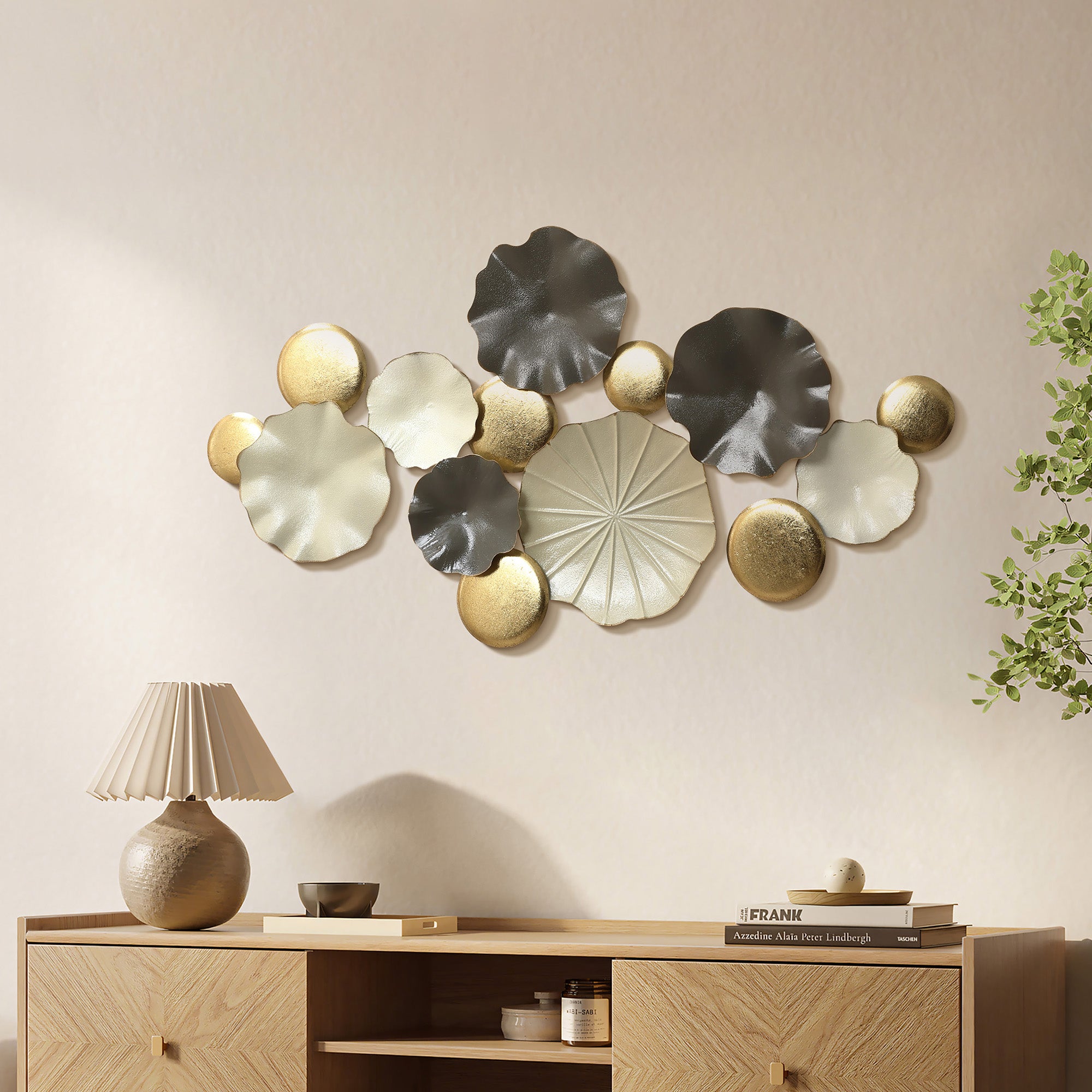 metal wall art decor with circular gold, white, and gray shapes hanging above a light wood sideboard with books, lamp, and decorative items, leafy plant on the right