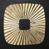 Square gold metallic object with a central square cutout and radiating, textured lines evokes striking sunburst wall art.
