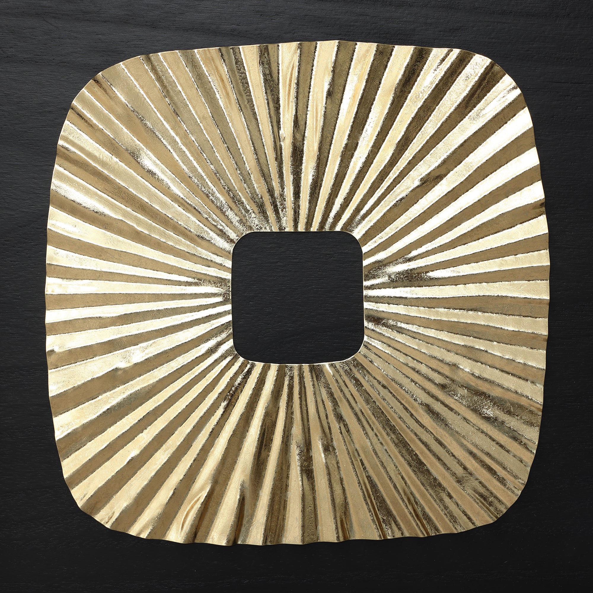 Square gold metallic object with a central square cutout and radiating, textured lines evokes striking sunburst wall art.
