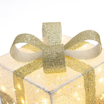 Square gift box wrapped in textured white paper with lights, topped with large glittery gold ribbon,bow.