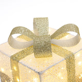 Square gift box wrapped in textured white paper with lights, topped with large glittery gold ribbon,bow.