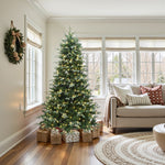 Pre-lit christmas tree with lights stands beside sofa in living room, with wrapped gifts at its base, festive wreath on the wall.