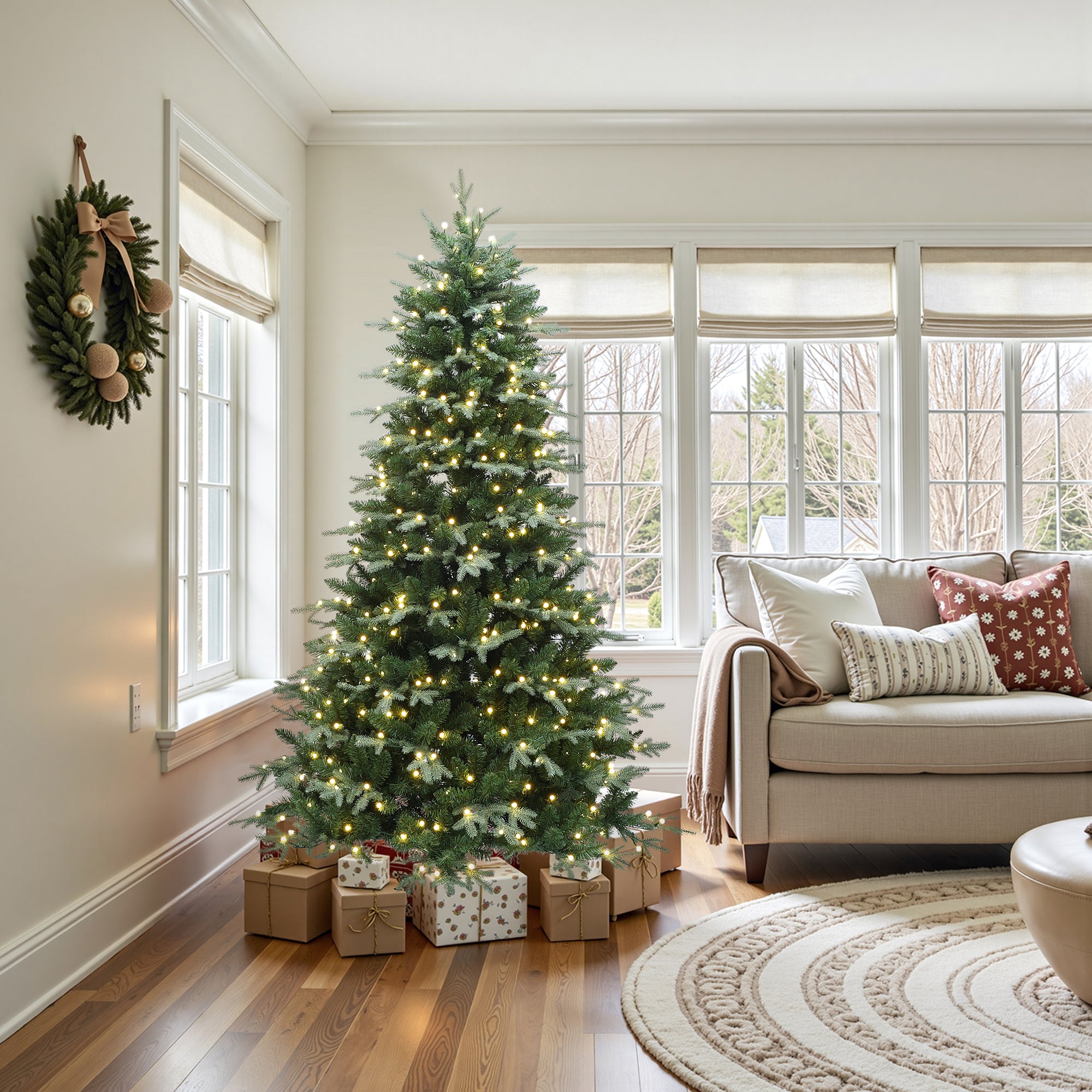 Pre-lit christmas tree with lights stands beside sofa in living room, with wrapped gifts at its base, festive wreath on the wall.