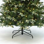 Pre-lit christmas tree with warm white string lights, supported by black metal stand on white surface.