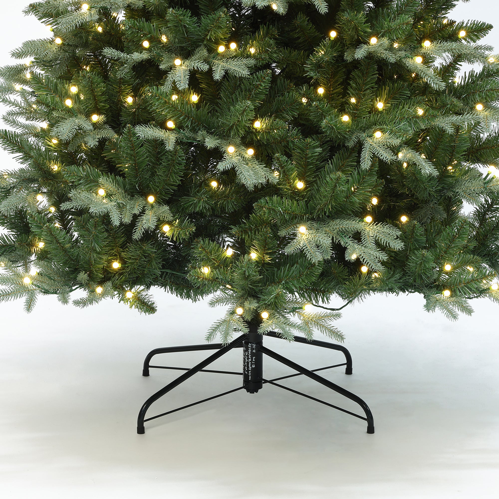 Pre-lit christmas tree with warm white string lights, supported by black metal stand on white surface.