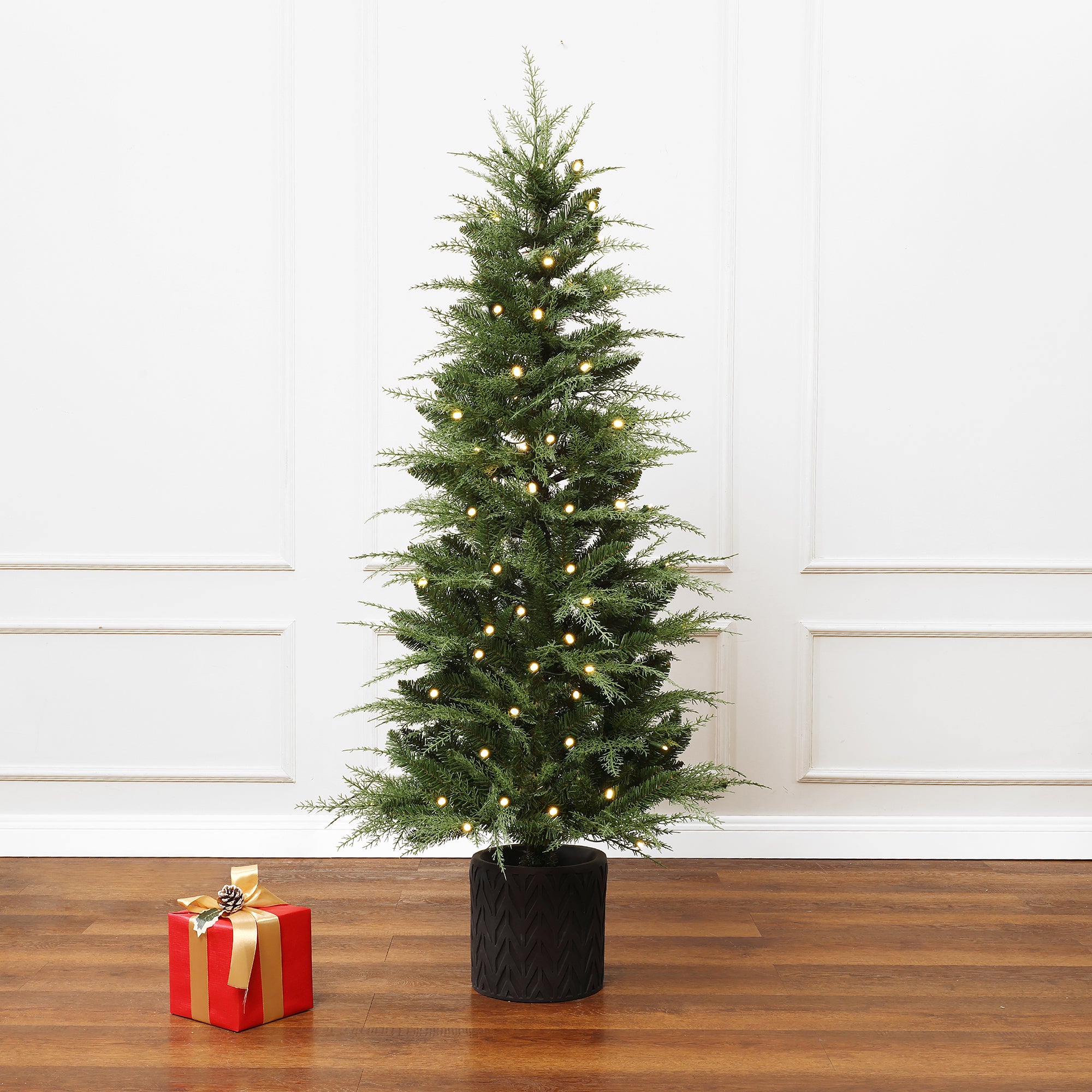 Decor 5ft artificial tree in black pot stands on wooden floor ,single red gift box with gold ribbon, sagainst white wall.
