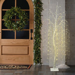 Wooden door with holiday wreath,garland sits beside glowing pre-lit christmas tree, white  string lights,against white brick wall.
