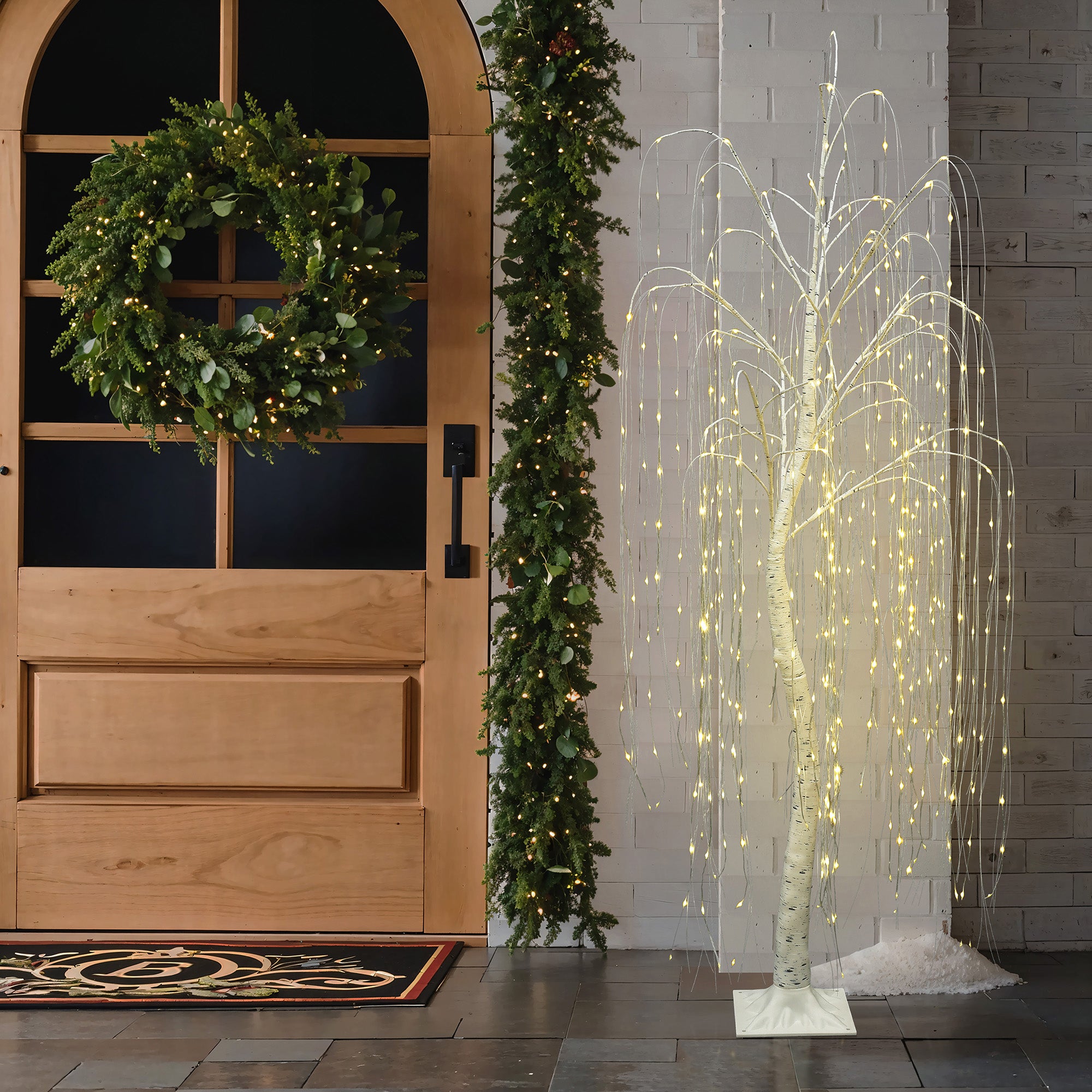 Wooden door with holiday wreath,garland sits beside glowing pre-lit christmas tree, white  string lights,against white brick wall.