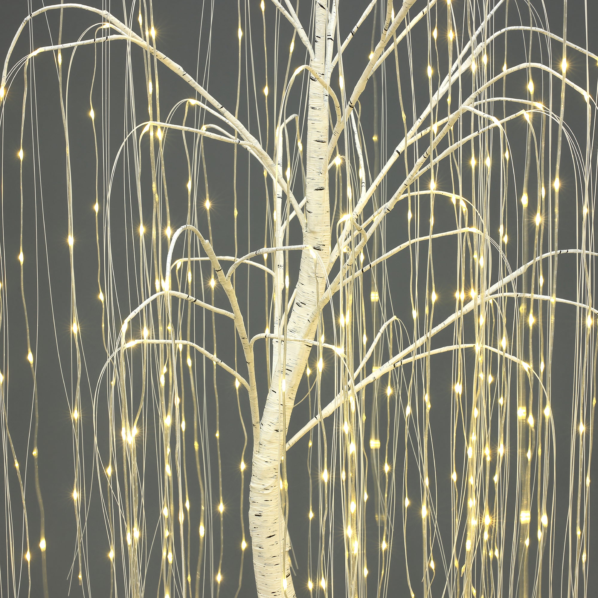 Close-up of artificial willow tree with thin branches, wrapped in small, warm white string lights against gray background.