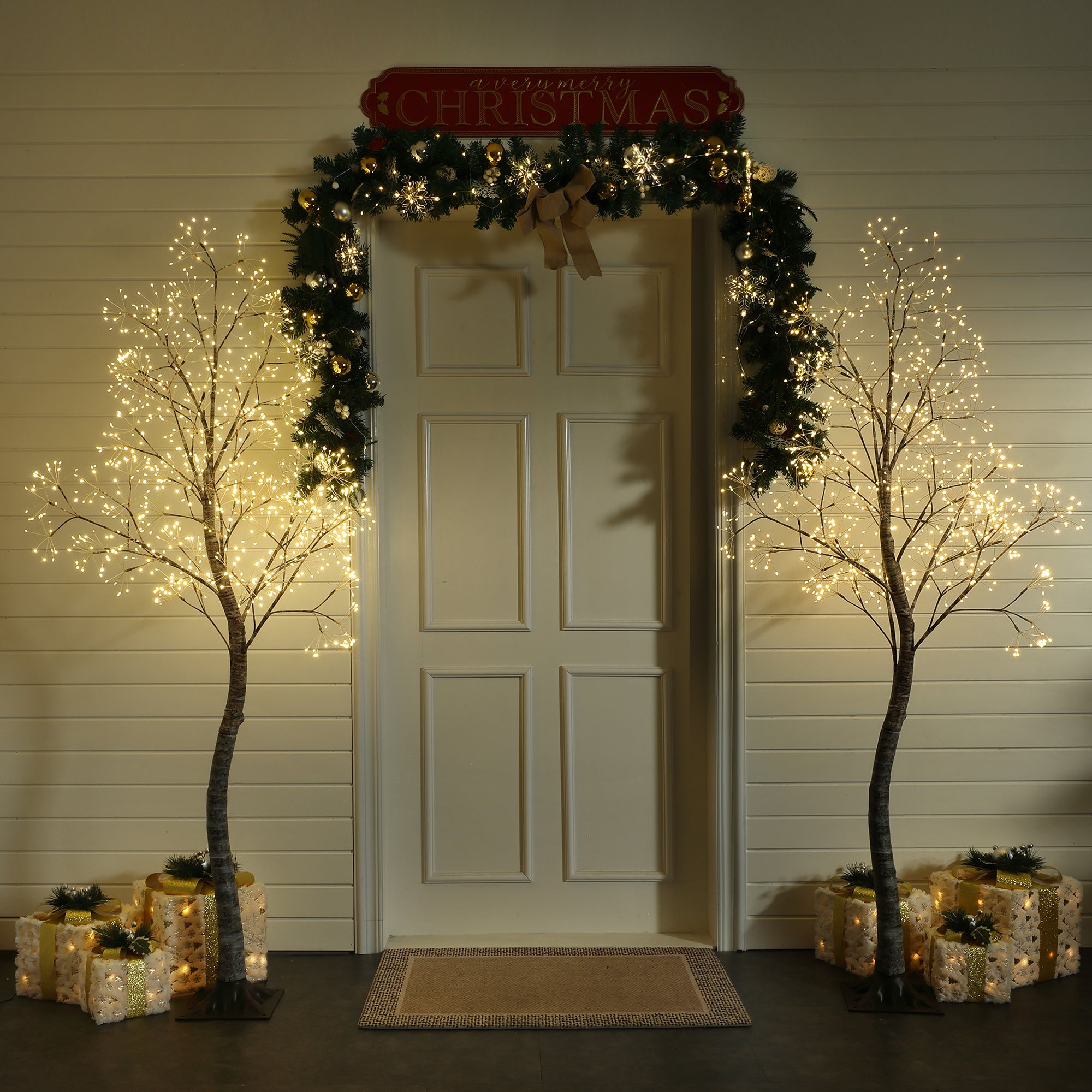 White door decor with christmas garland, 2 lit led trees on each side, wrapped presents arranged around the base.
