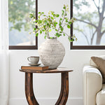 Textured cement vase holding green branches, a stacked book, set beside a white sofa in front of large windows.