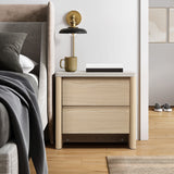 Light wood nightstand with two drawers sits beside a bed, topped with a lamp, mug, and books.