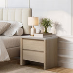 Light wood modern end table with two drawers sits beside a bed, topped with a lamp, a small plant, stacked books.