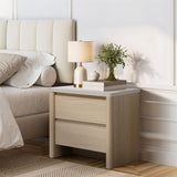 Light wood modern end table with two drawers sits beside a bed, topped with a lamp, a small plant, stacked books.