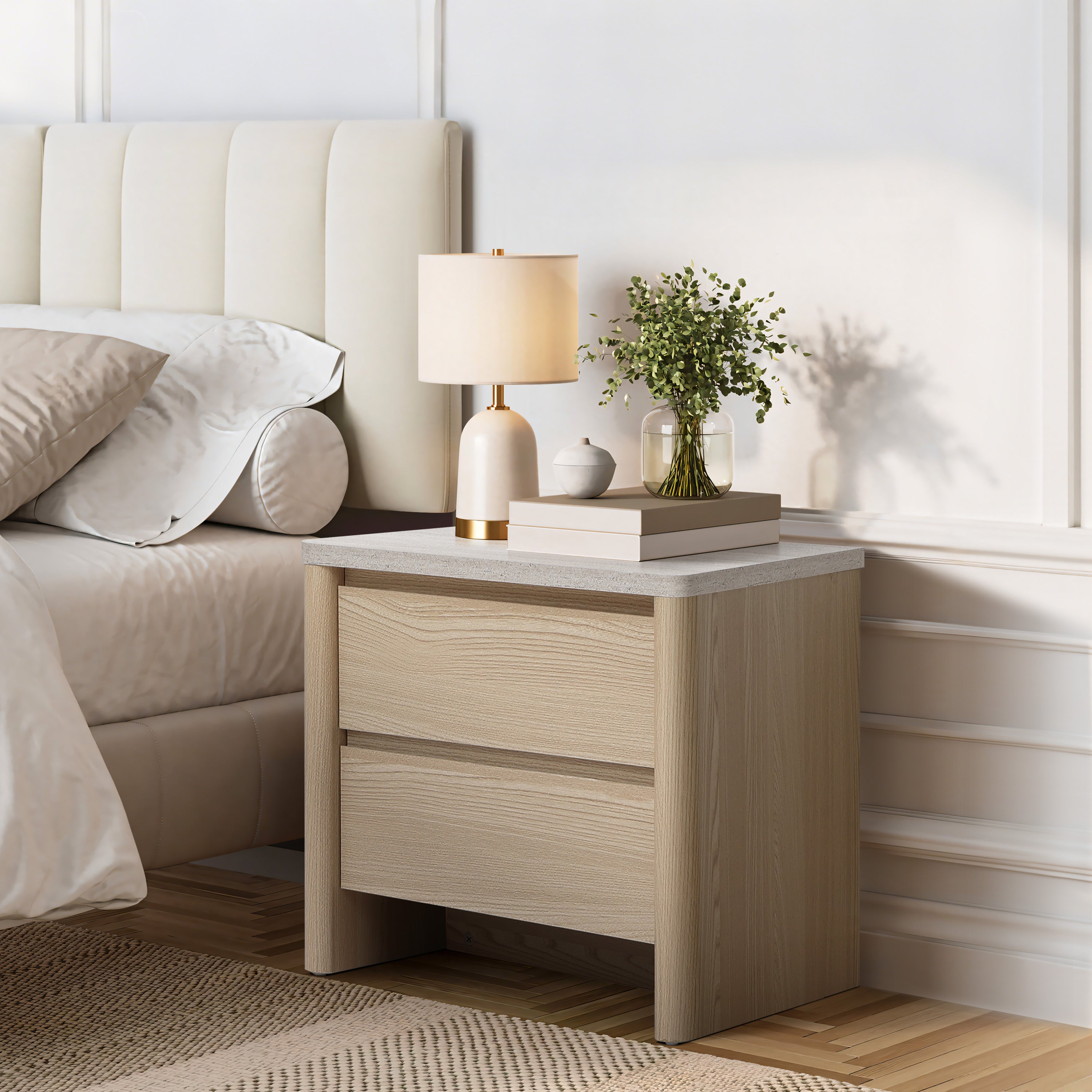 Light wood modern end table with two drawers sits beside a bed, topped with a lamp, a small plant, stacked books.