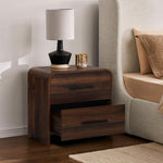 A walnut nightstand with two drawers, one partially open, holds a black,beige lamp,a gold candle.