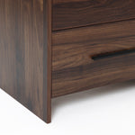 Close-up of the lower corner of a walnut nightstand with a dark wood grain finish,a sleek horizontal black handle.