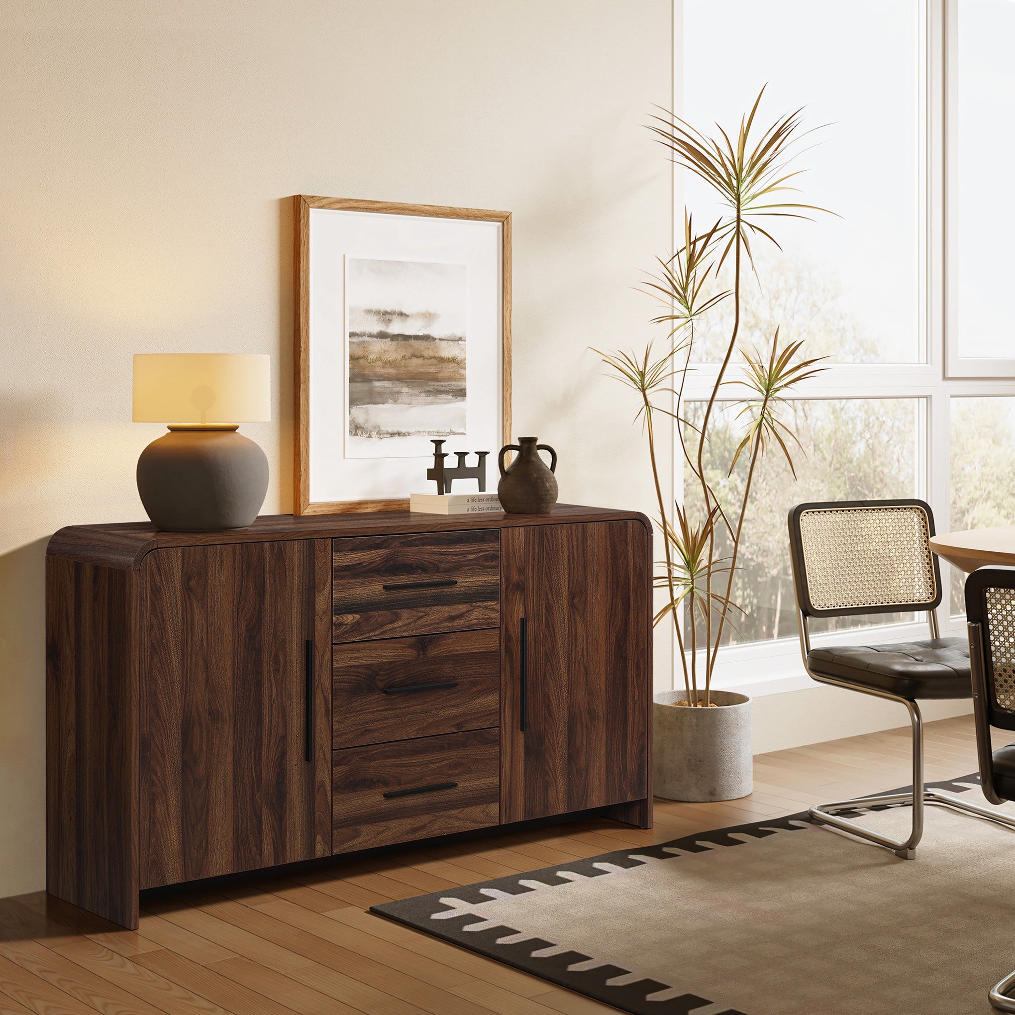 A wood sideboard cabinet with a lamp, framed art, decor items beside a potted plant, chair.