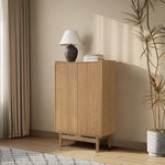 Modern light oak wood storage cabinet stands against a beige wall beside a tall plant and a window with curtains.