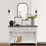 A farmhouse-inspired table with four drawers features a black lamp, framed art, a vase with greenery, and a bowl. A basket sits on the lower shelf, while a mirror and two sconces hang above this charming storage console table.