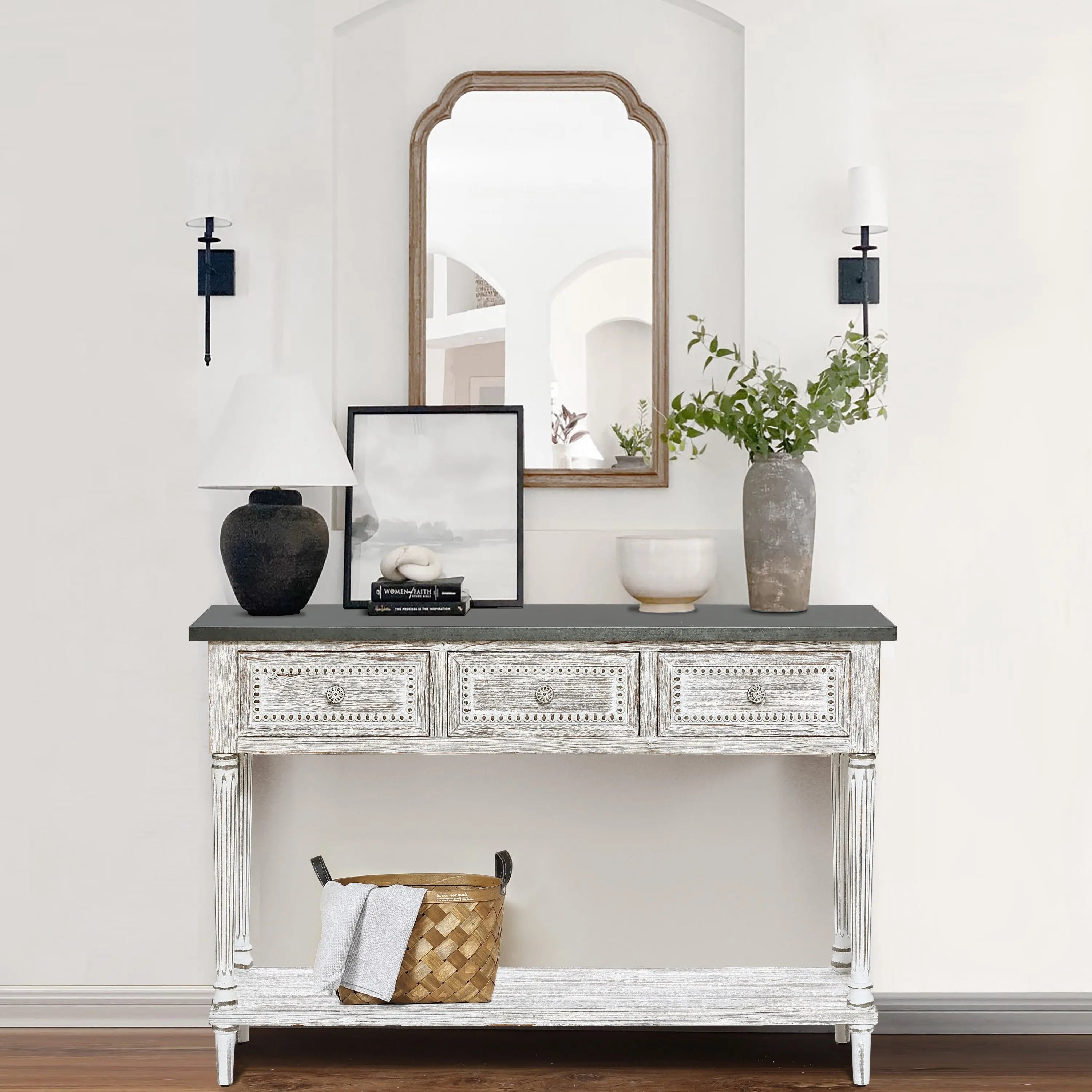 A farmhouse-inspired table with four drawers features a black lamp, framed art, a vase with greenery, and a bowl. A basket sits on the lower shelf, while a mirror and two sconces hang above this charming storage console table.