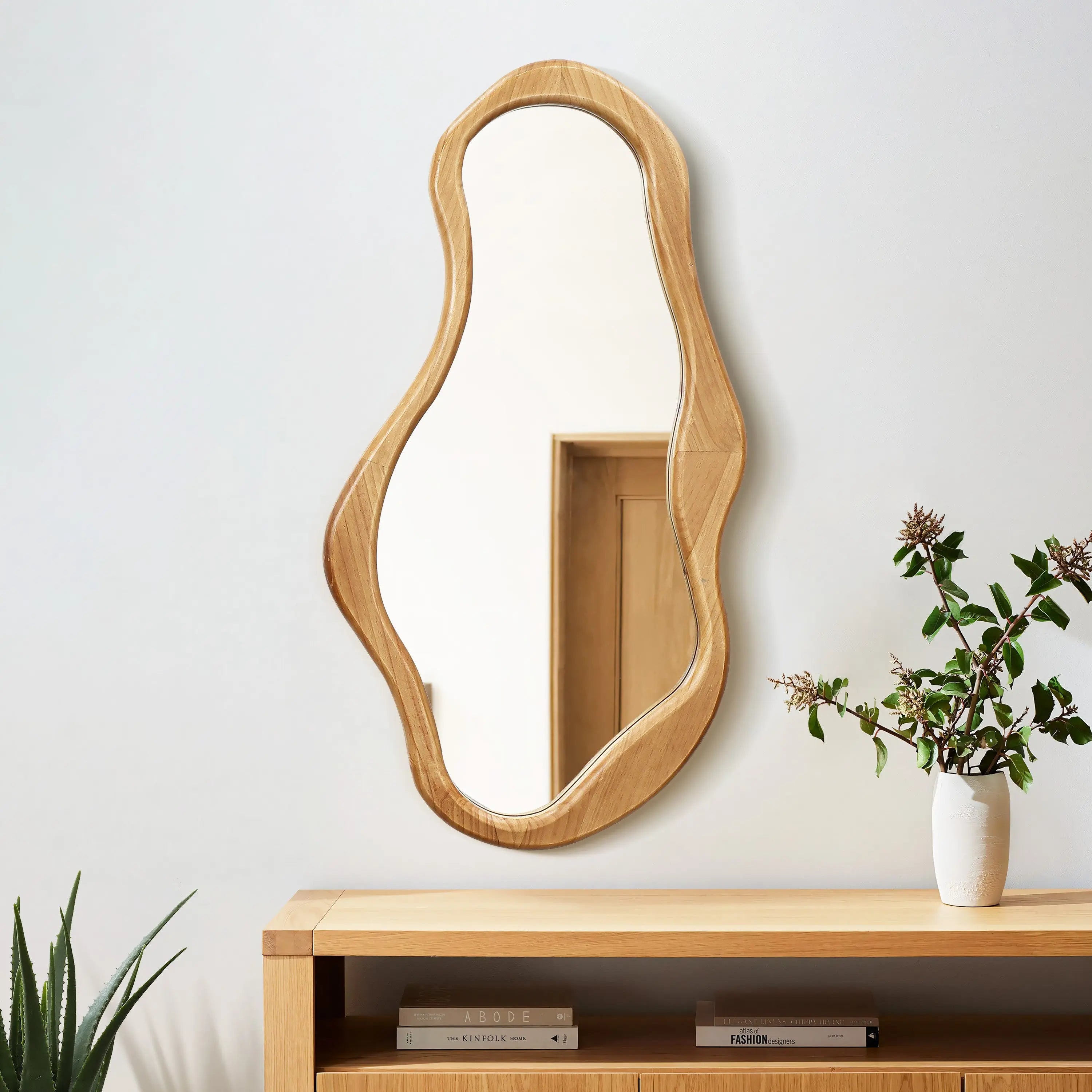 An irregularly shaped mirror with a pine frame hangs on a wall above a wooden console, infusing the space with natural energy. A potted plant with green and brown leaves sits on the console, enhancing the organic decor.