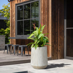 A tall, striped magnesium oxide planter filled with lush green tropical plants adorns the patio near a wooden building with large windows and sleek black bar stools.