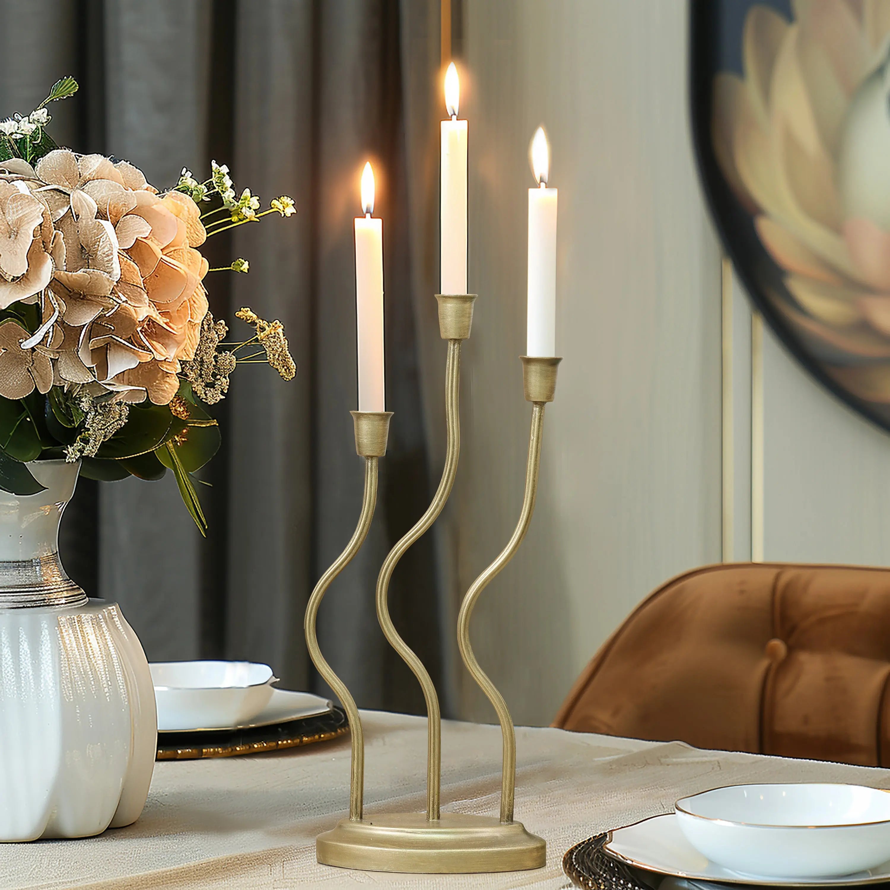 A three-branch candle holder with an elegant design sits on the table, its lit candles casting a warm glow beside a vase of beige flowers, with plates and a chair subtly enhancing the homes decor in the background.