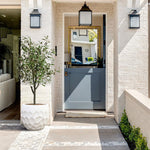 A white brick house entrance with a gray Dutch door, flanked by black lanterns and a geometric pot planter. The planter, crafted from durable magnesium oxide material for indoor and outdoor use, stands beside a small rug on the step. Another house is reflected in the door window.
