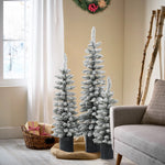 Three flocked artificial Christmas trees with PVC branches stand in a living room corner near a window, a wicker basket of gifts, and a gray armchair. A wreath hangs on the beige wall.
