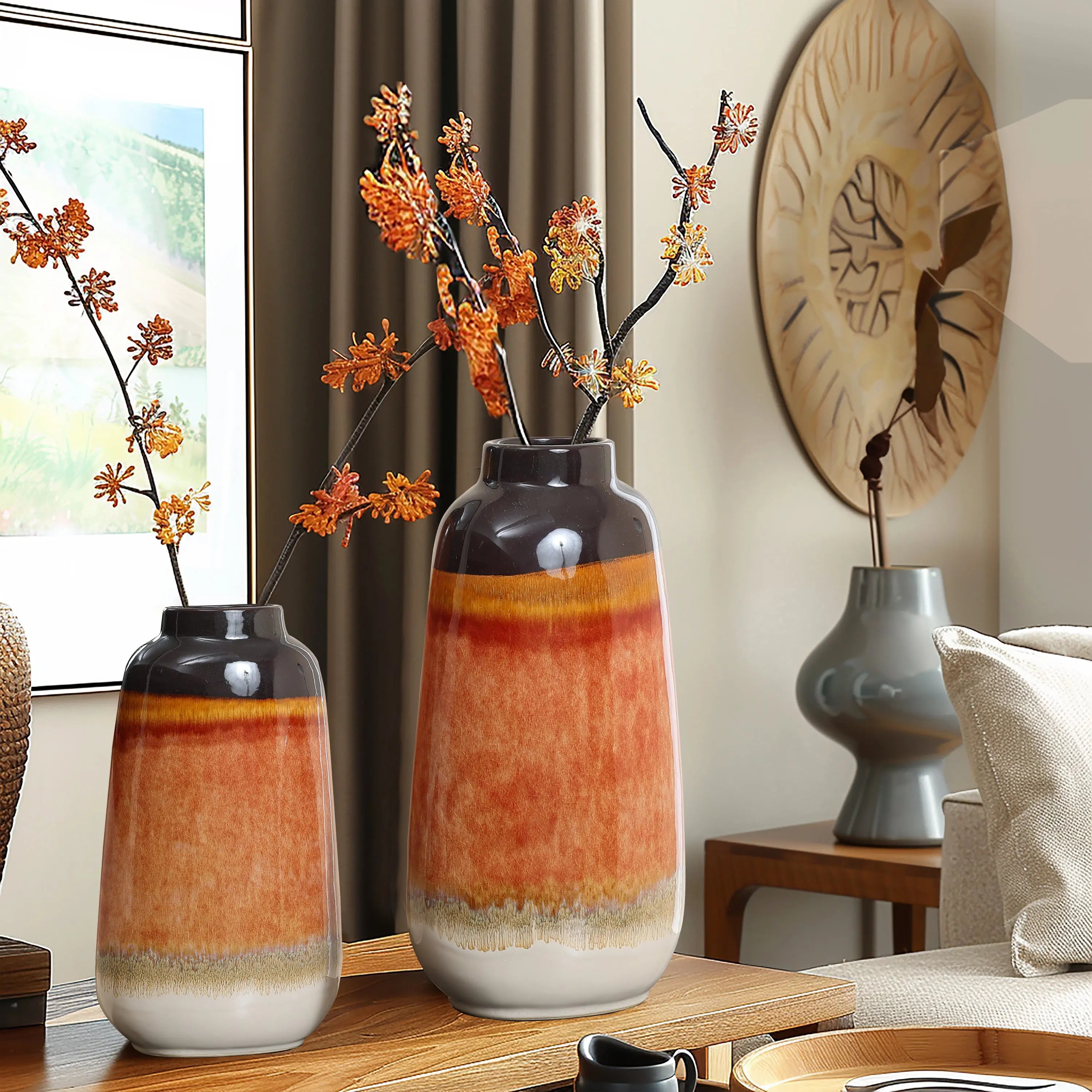Two decorative vases with orange and gray hues, featuring a color block gradient, contain branches with orange blossoms. They rest on a wooden table in a modern, neutral-toned room, adding charm to your homes decor.